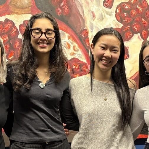 Three alums and a teacher stand in front of a painting of a pomegranate.