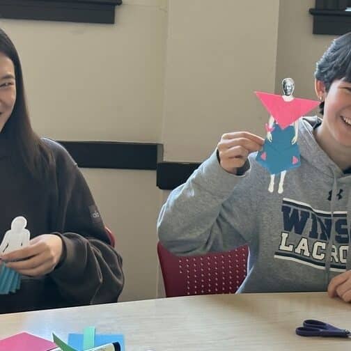 Two students work on zero-waste fashion and hold up paper dolls.