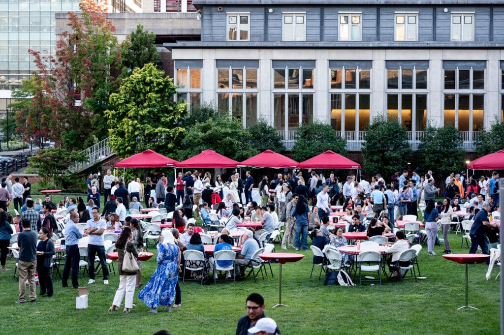 People socializing outside in a grassy courtyard standing and seated at tables
