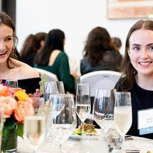 Two alums sit at a table having dinner in a crowded room.