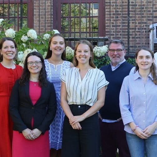 Nine adults stand outside in front of flowering bushes
