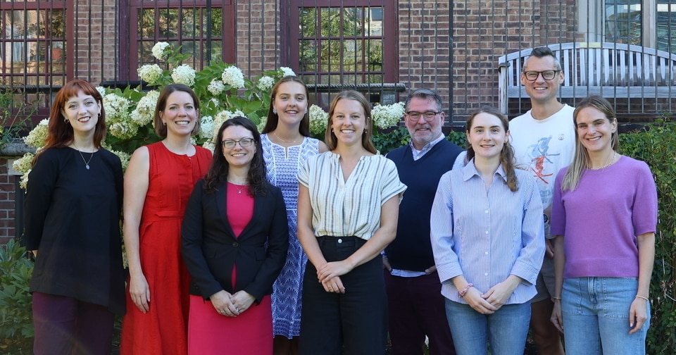 Nine adults stand outside in front of flowering bushes