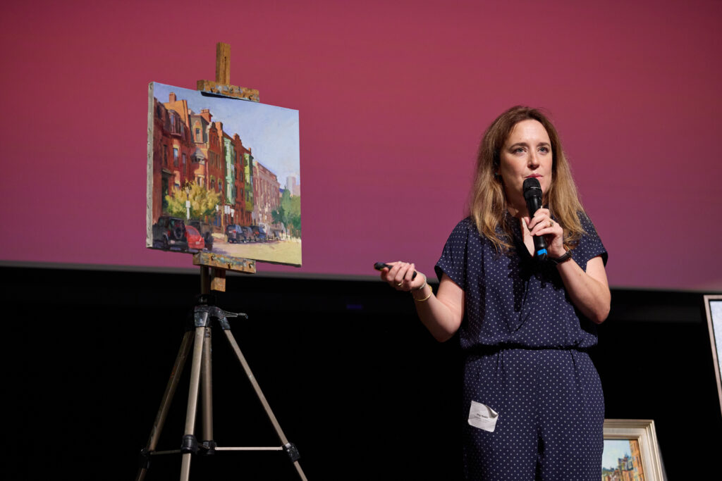 Woman with long brown hair in a blue jumpsuit, standing in front of an easel holding a painting of buildings.