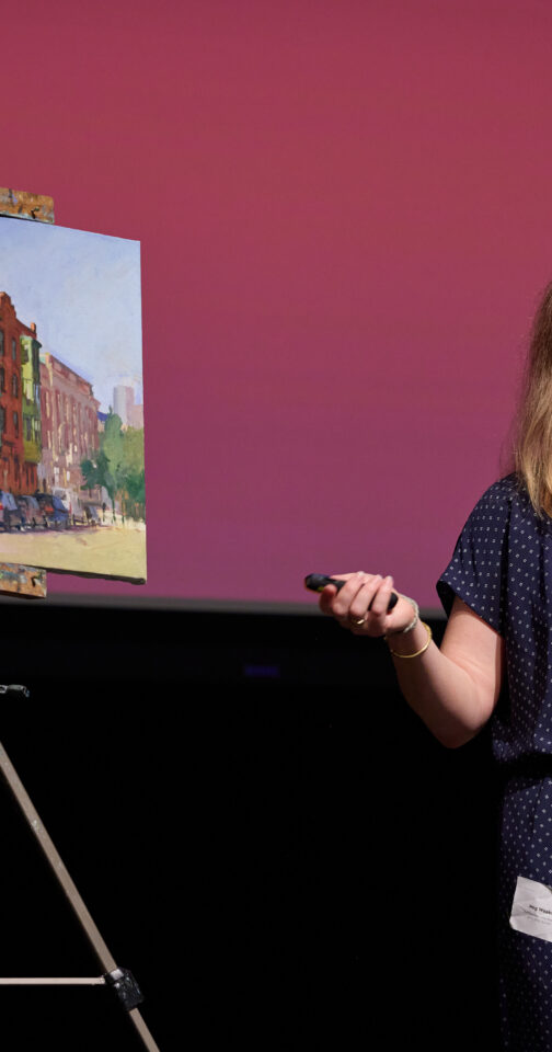 Woman with long brown hair in a blue jumpsuit, standing in front of an easel holding a painting of buildings.