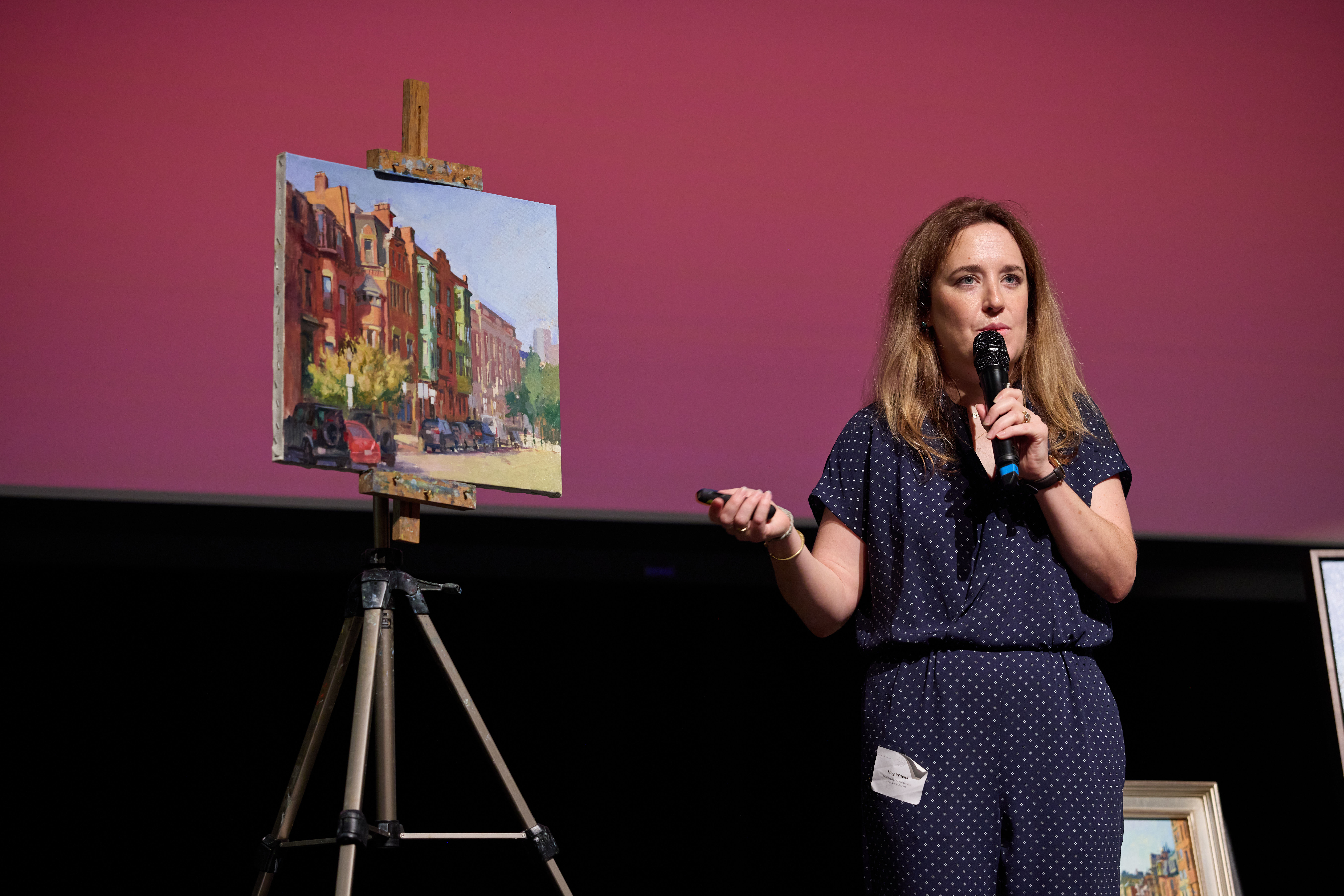Woman with long brown hair in a blue jumpsuit, standing in front of an easel holding a painting of buildings.
