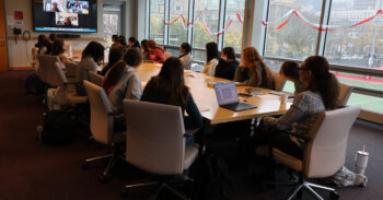 students and a teacher seated around a conference table, facing a television with others on the screen for a remote discussion