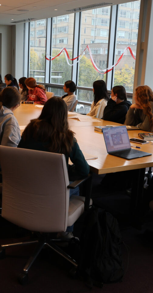students and a teacher seated around a conference table, facing a television with others on the screen for a remote discussion
