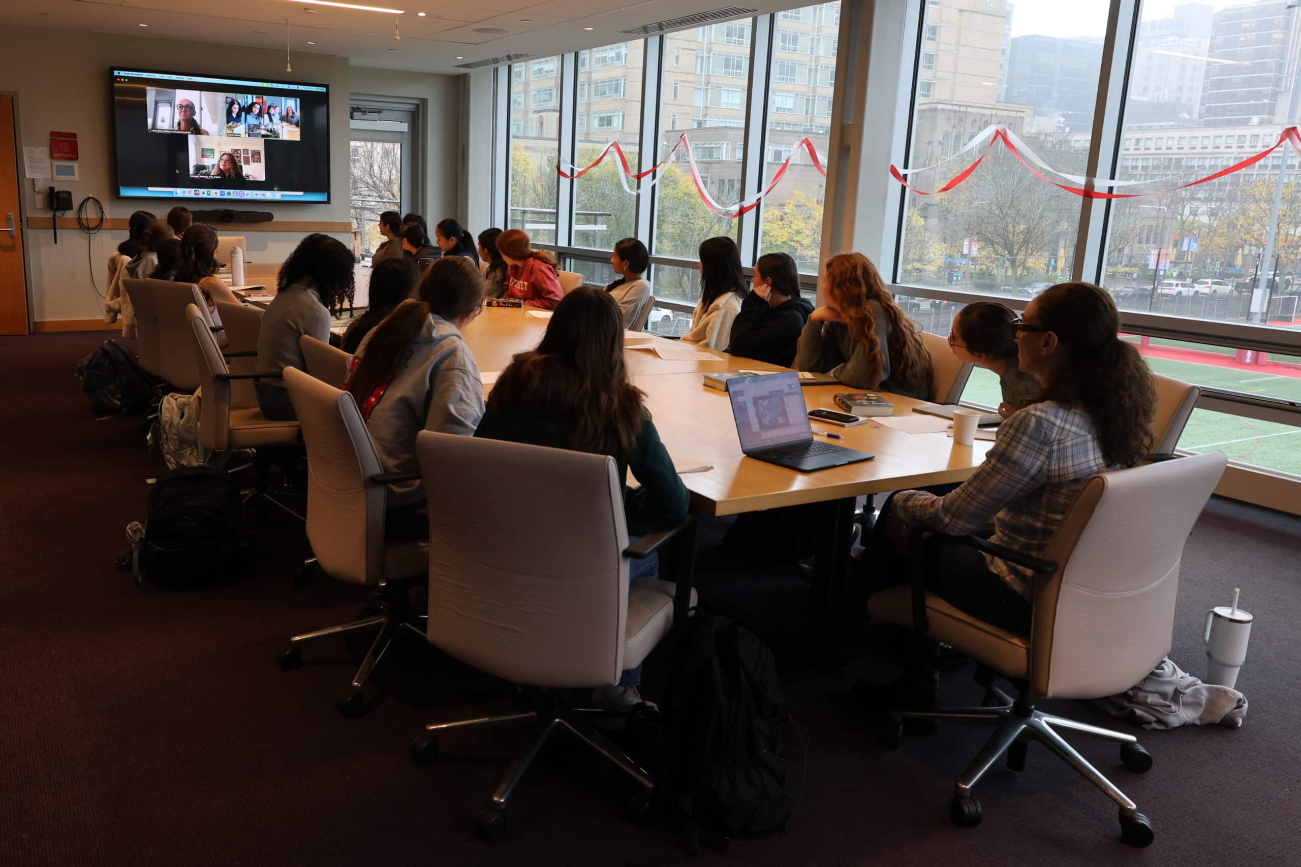 students and a teacher seated around a conference table, facing a television with others on the screen for a remote discussion