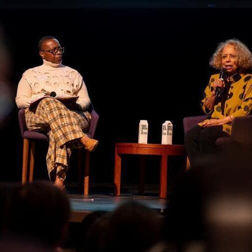 Two women of color seated on a stage holding microphones.