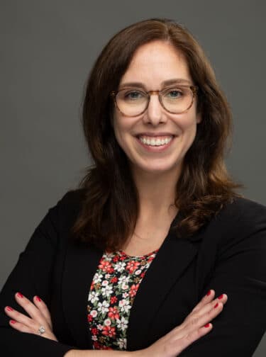 White woman with dark hair and glasses, wearing a blazer, arms crossed